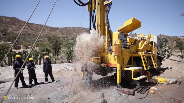 Still from a charity: water video: a yellow drilling rig blasts water and rock into the air while a crew in hard hats looks on.