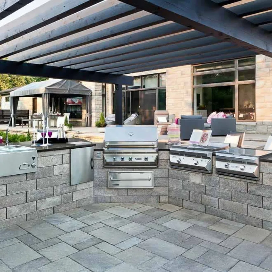 An outdoor cooking area built from stone pavers, with a built-in grill, counter seating and a dining set on a patio.