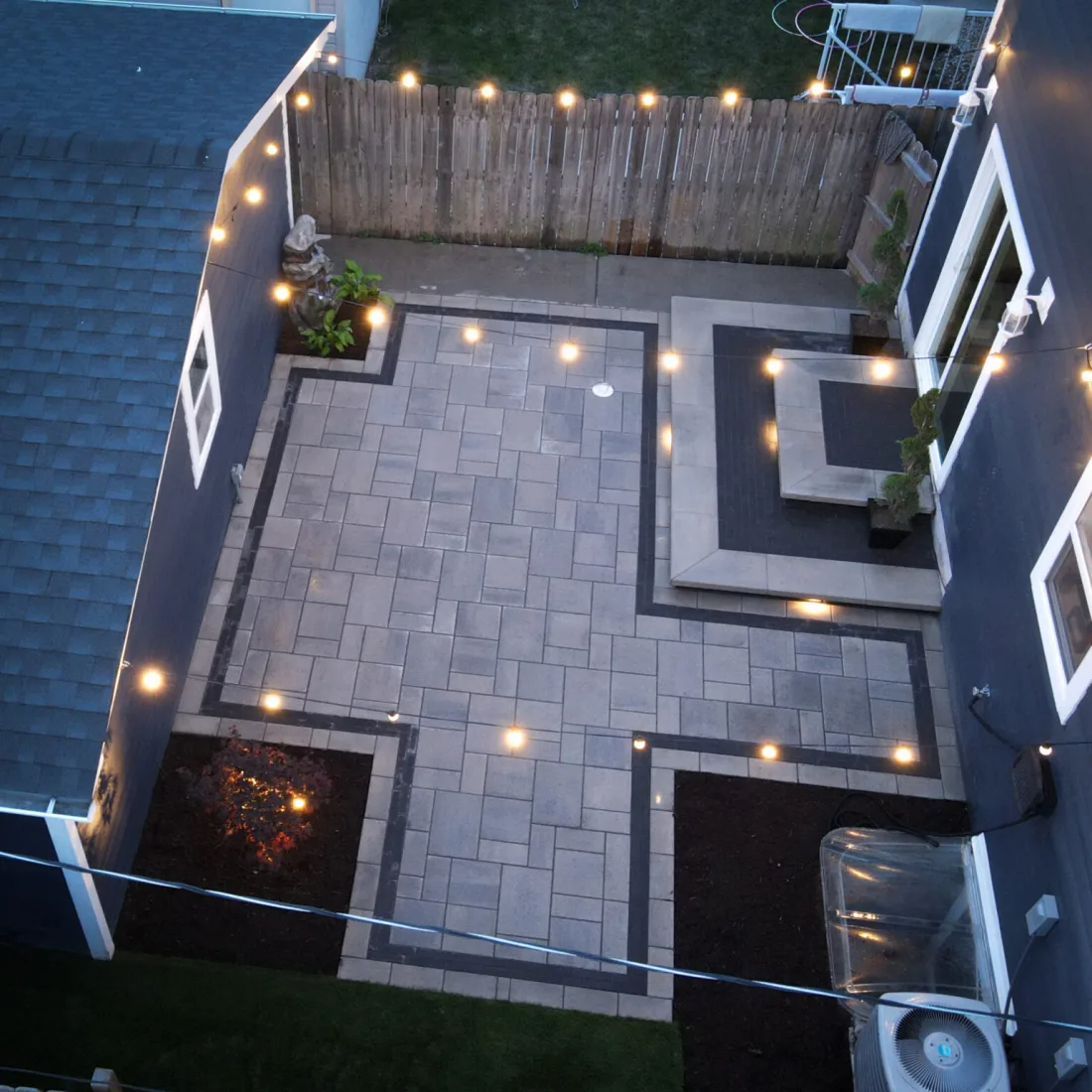 The same backyard after the build: a grey and charcoal paver patio with a raised seating area, planting beds and low-voltage lighting at dusk.